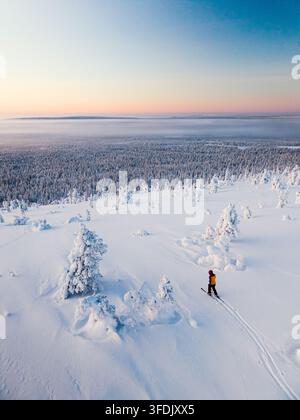 Vue aérienne d'un fondeur glissant à travers un vaste paysage forestier enneigé sous un ciel crépusculaire doux à Muonio, en Laponie, en Finlande. Banque D'Images