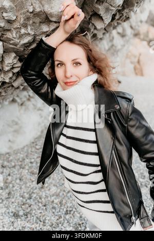 Veste en cuir pour femme, brune attirante portant un pull rayé et une veste moto noire, appuyée contre une paroi rocheuse côtière Banque D'Images