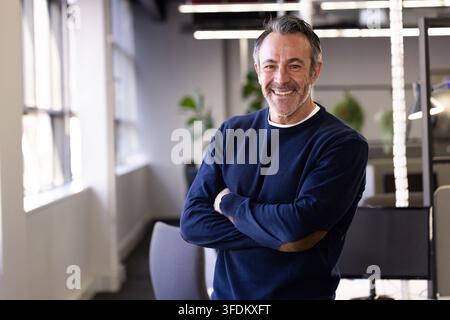 Homme senior debout, les bras croisés et souriant dans un bureau moderne avec des bureaux et des écrans Banque D'Images