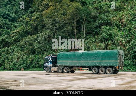 Ngoc Linh, Vietnam - 22 août 2026 : Un grand camion de marchandises avec une remorque couverte est garé sur un pavé mouillé à côté d'une forêt tropicale dense. Banque D'Images