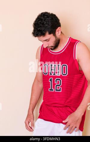 Homme barbu en débardeur rouge, la tête baissée, déçu par le résultat du match ; il porte un short blanc. Banque D'Images