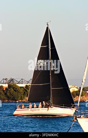 Un voilier de course avec des voiles noires et un équipage assis traverse l'eau bleue près du rivage de San Diego sous la lumière chaude de la fin d'après-midi. Banque D'Images
