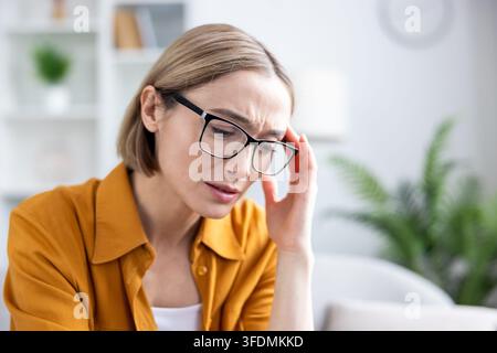 Femme portant des lunettes ressentant une forte douleur et un inconfort dus à un mal de tête, une migraine ou du stress, touchant sa tempe tout en souffrant à la maison ou au bureau Banque D'Images