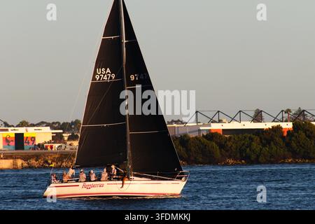 Un voilier de course avec des voiles noires et un équipage assis traverse l'eau bleue près du rivage de San Diego sous la lumière chaude de la fin d'après-midi. Banque D'Images