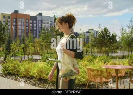 Une femme traverse le parc en tenant un sac en filet réutilisable rempli de légumes. Des arbres verts et des bâtiments se trouvent en arrière-plan Banque D'Images