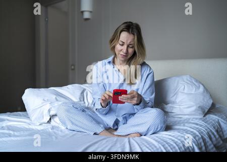 Femme d'âge moyen fatiguée assise sur le lit en pyjama se sent épuisée tout en utilisant son téléphone au lit après s'être réveillée. Nuit d'insomnie, addiction aux écrans. Épuisée Banque D'Images