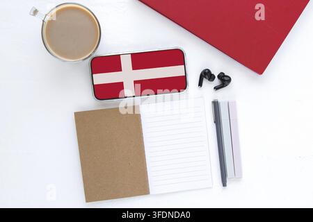 Smartphone avec le drapeau du Danemark sur l'écran, ordinateur portable ouvert, tasse de café et papeterie sur une table blanche, concept d'apprentissage des langues en ligne, éducation et st Banque D'Images