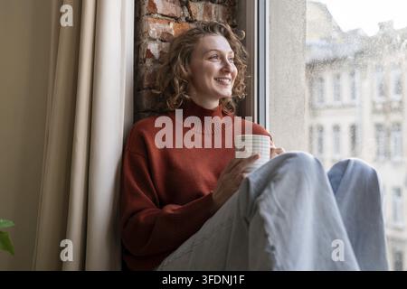 Femme heureuse profitant d'un moment confortable avec une boisson chaude par fenêtre. Femme souriante se sentant détendue, prend une pause des tâches quotidiennes avec une tasse de café. Mindfulnes Banque D'Images
