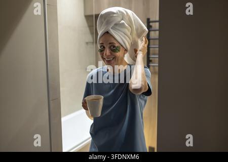 Femme d'âge moyen souriante et joyeuse sort de la salle de bain avec une serviette sur la tête, des patchs oculaires, profitant de sa routine matinale avec une tasse de thé. Ambiance bien-être d'une personne heureuse Banque D'Images