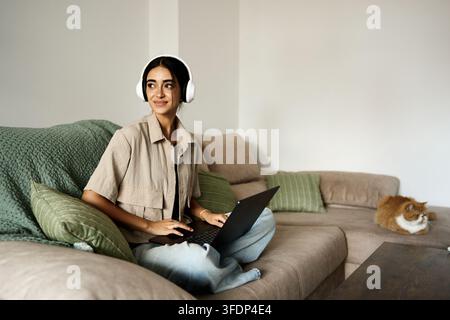 Jeune femme portant des écouteurs et tapant sur un ordinateur portable, assise confortablement sur un canapé à la maison avec son chat. Concept de travail à distance Banque D'Images