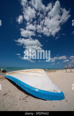 Bateau de pêche blanc et bleu renversé reposant sur une plage de sable tranquille sous des nuages blancs dramatiques et un ciel bleu profond. Banque D'Images
