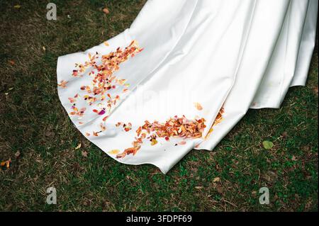 La traîne de la robe de mariée blanche de la mariée posée sur l'herbe verte parsemée de pétales de fleurs. Banque D'Images