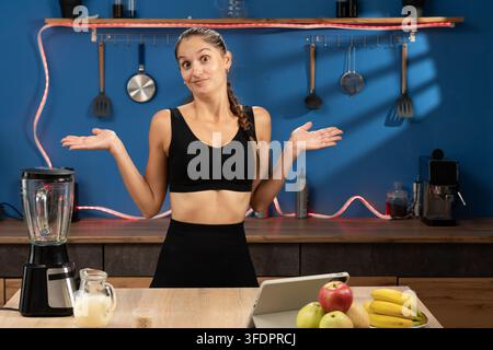 Un blog santé. Une jeune blogueuse fitness en tenue de sport se tient dans sa cuisine avec un mur bleu, regardant la caméra et écartant les bras Banque D'Images