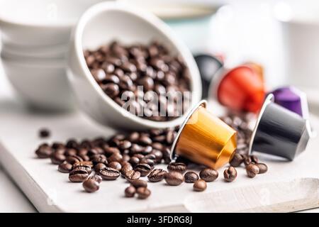 Close-up d'une capsule de café dorée nichée dans un bol de grains de café torréfiés noirs avec des capsules et des grains éparpillés sur une surface blanche, évoquant un arôme frais Banque D'Images