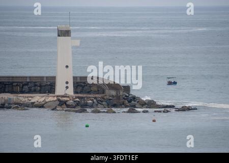 Phare solitaire se dressant sur une jetée rocheuse du port tandis qu'un petit bateau de pêche couvert dérive au large ; une mer bleu-gris atténuée et un ciel nuageux créent une qu Banque D'Images