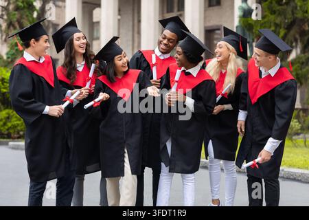 Diplômés heureux, groupe international de jeunes hommes et femmes en costumes de graduation tenant des diplômes, se regardant et discutant, ayant cérémonie Banque D'Images