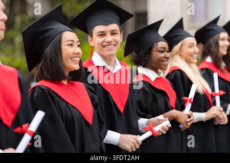 Jeune homme charmant et souriant debout parmi un groupe international d'étudiants, portant des costumes de remise des diplômes et tenant des diplômes, des diplômés multiraciaux ayant g Banque D'Images