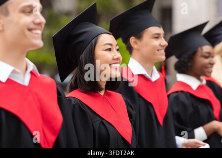 Groupe multiculturel d'étudiants joyeux lors d'une cérémonie de remise des diplômes, portant des robes et des toges noires et rouges, debout en rang à l'université Banque D'Images