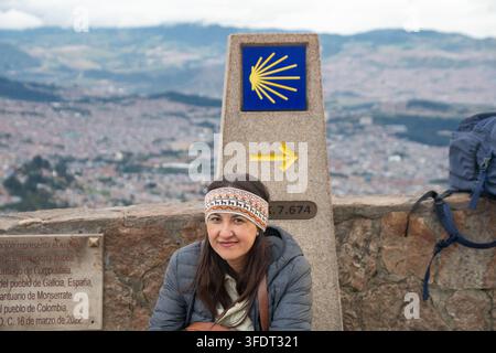 Une pèlerine souriante est assise près d'un marqueur en coquille de Saint-Jacques et d'une flèche jaune du Camino de Santiago au sanctuaire de Monserrate, surplombant Bogotá, Colombie Banque D'Images