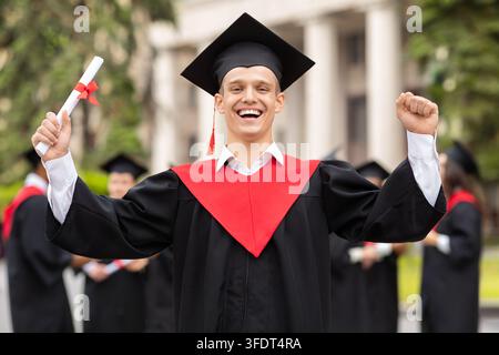 Étudiant ravi organisant une fête de fin d'études, portant une robe et un chapeau de diplôme, tenant un diplôme et levant les mains en l'air, posant devant un groupe international Banque D'Images