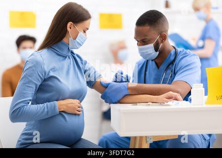 Médecin afro-américaine administrant une injection de vaccin contre le coronavirus à une femme enceinte, portant un masque de protection dans une clinique d'hôpital. Vaccin contre le virus Corona Banque D'Images