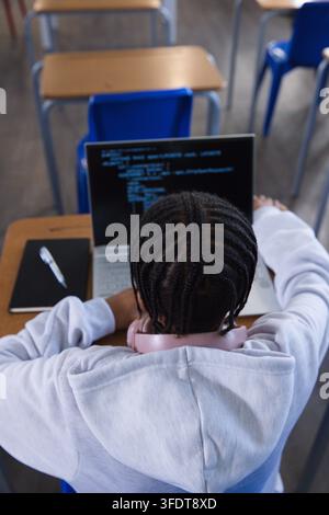 Étudiante afro-américaine tapant du code sur un ordinateur portable à un bureau en bois, cahier, stylo, casque rose Banque D'Images
