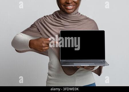 Femme africaine dans hijab pointant vers ordinateur portable avec écran noir, Mockup image Banque D'Images