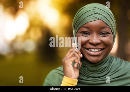 Portrait de la gaie noire musulmane dans hijab parlant dessus Téléphone portable extérieur Banque D'Images