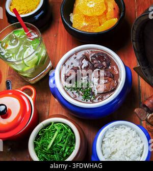 Feijoada brésilienne traditionnelle avec du riz, des choux verts, de la farofa, des tranches d'orange et un caipirinha au citron vert sur une table en bois rustique. Banque D'Images