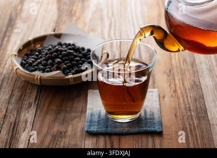 Verser du thé de soja noir d'une théière en verre dans un verre posé sur un dessous de verre Banque D'Images