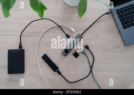 Hub USB en aluminium reliant un ordinateur portable à un disque dur externe, un SSD portable, une carte mémoire et des câbles sur un bureau en bois. Expansion de ports et emplacements supplémentaires Banque D'Images
