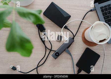 Câbles USB emmêlés, un hub de docking, des disques durs externes et une carte mémoire encombrent un bureau en bois à côté d'un ordinateur portable et d'une tasse de café. Espace de travail désordonné pour Banque D'Images
