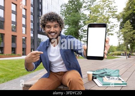 Jeune homme souriant et assis en plein air sur un campus universitaire, tenant un smartphone avec un écran blanc vierge et y pointant, présentant une nouvelle technologie mobile Banque D'Images