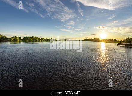 Le soleil doré plonge bas au-dessus de la surface calme de la rivière. Le ciel brille d’un bleu parsemé de nuages doux au-dessus. Les arbres bordent la rive lointaine Banque D'Images