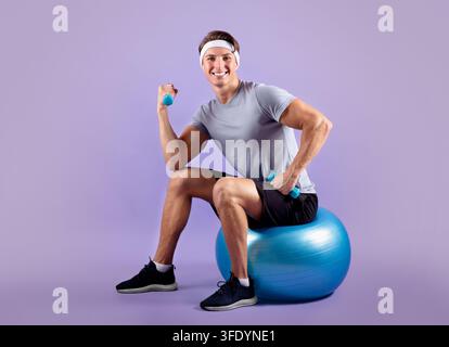 Jeune homme en vêtements de sport assis sur un ballon de fitness et soulevant des haltères Banque D'Images