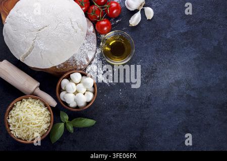 Ingrédients de la pizza margherita pâte crue tomates cerises basilic huile d'olive fromage de mozzarella sur une table de cuisine en pierre sombre Banque D'Images
