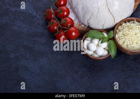 Ingrédients de la pizza margherita pâte crue tomates cerises basilic huile d'olive fromage de mozzarella sur une table de cuisine en pierre sombre Banque D'Images