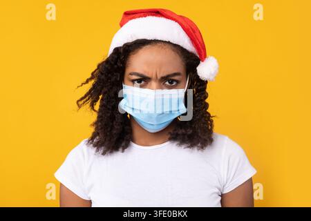 Femme africaine malheureuse portant un masque et un chapeau de Noël, fond jaune Banque D'Images