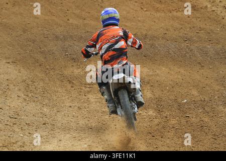 Vue arrière d'un pilote de motocross vêtu d'un équipement orange vif et d'un casque bleu, soulevant de la poussière sur une piste en terre battue lors d'une épreuve de course tout-terrain. Banque D'Images