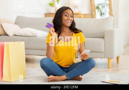 Concept de cybershopping. Femme afro-américaine avec smartphone et carte de crédit commandant des choses en ligne à la maison Banque D'Images