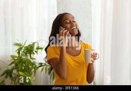 Charmante femme noire parlant au téléphone portable avec une tasse d'arôme café à la maison Banque D'Images