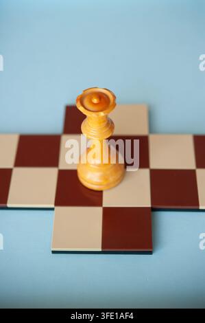 Close-up d'une dame d'échecs en bois clair posée sur une petite croix d'échecs rouge et crème devant un fond bleu pâle Banque D'Images
