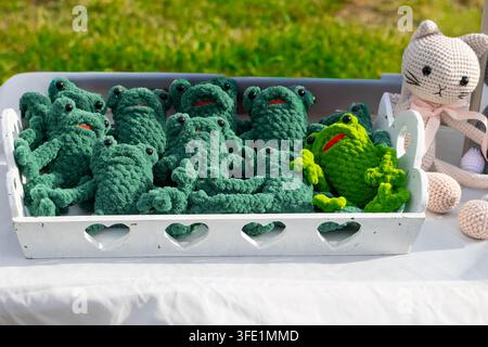 Collection de jouets en peluche tricotés à la main en forme de grenouille disposés dans un plateau en bois blanc décoratif sur un marché artisanal en plein air. Sélection artisanale comprenant gr Banque D'Images