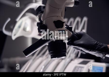 Homme concentré, 30 ans, ajuste un microscope dans une clinique dentaire stérile. Un gant noir sur la main met en valeur la précision dans le travail scientifique parmi les instruments dentaires Banque D'Images