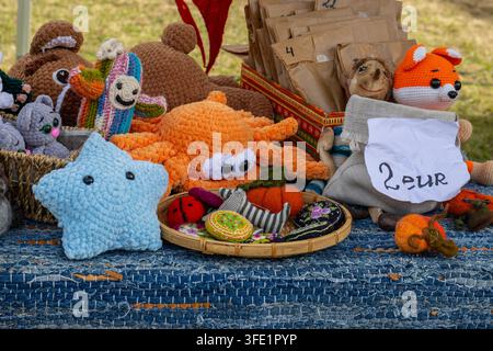 Collection colorée de jouets en peluche amigurumi crochetés à la main et d'accessoires feutrés disposés sur une nappe bleue tissée à un marché artisanal en plein air Banque D'Images