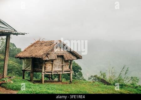 Une maison traditionnelle sur pilotis en bambou avec un toit de chaume se dresse sur une pente herbeuse d'une montagne au-dessus d'une vallée brumeuse. Banque D'Images