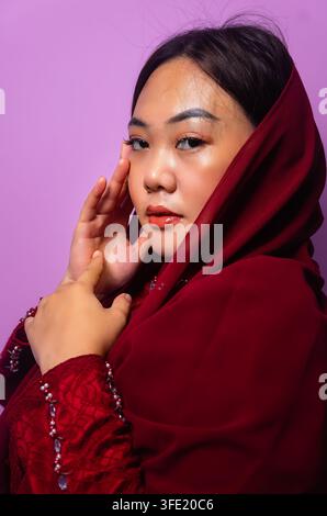belle femme asiatique portant un hijab rouge et une tenue traditionnelle, posant doucement contre un fond violet, prise en studio. Banque D'Images