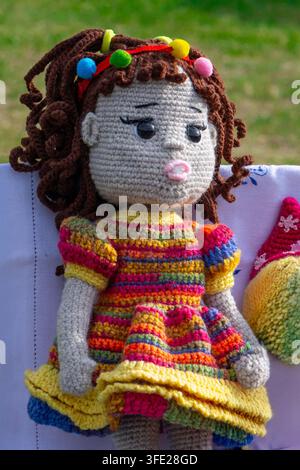 Portrait rapproché d'une poupée amigurumi tricotée à la main avec des cheveux bouclés bruns décorée de pompons colorés, portant un st vibrant multicolore Banque D'Images