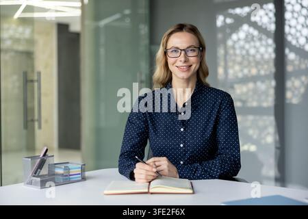 Femme d'affaires portant des lunettes souriant avec confiance tout en étant assise à son bureau, tenant un stylo et prenant des notes dans un cahier ouvert, démontrant un professionnalisme et une attitude de travail positive Banque D'Images