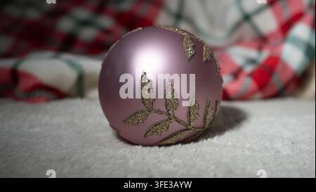 Close-up d'une boule de Noël rose avec un motif de branche de fougère pailletée or étincelant sur une couverture en peluche blanche, fond en tissu tartan rouge doux à carreaux Banque D'Images